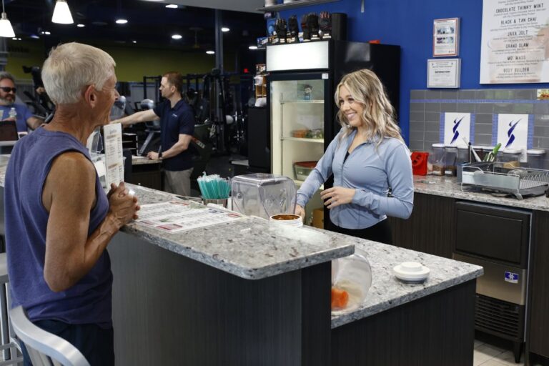 A gym member chats with a smiling fitness center employee as she prepares a shake at the onsite Shore Shakes bar in an Atlantic Highlands gym